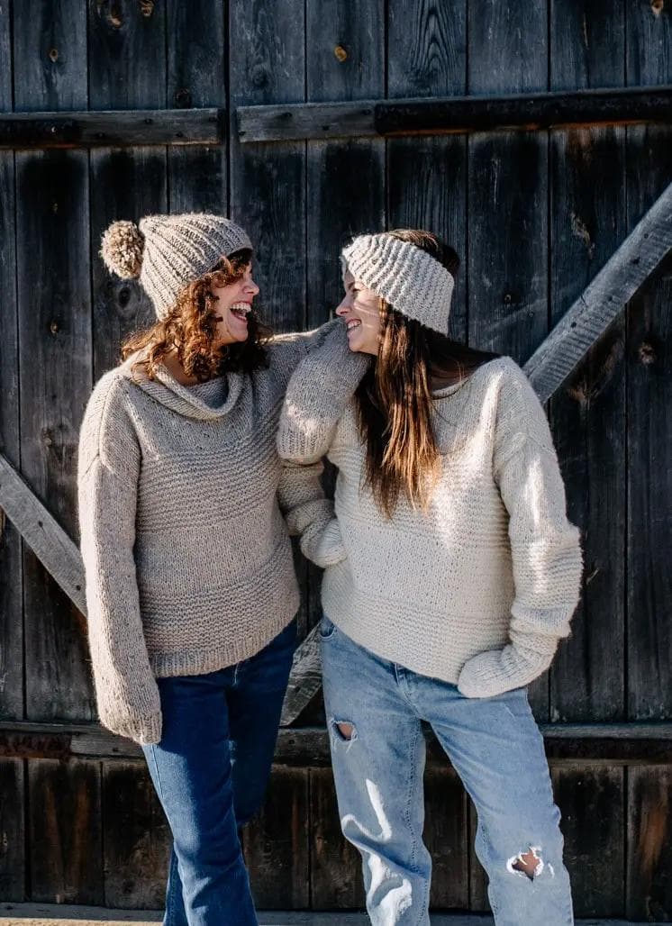 Na zdjęciu znajdują się dwie uśmiechnięte kobiety, które patrzą na siebie. Kobiety stoją przed drewnianymi drzwiami, obie maja na sobie grube swetry. Kobieta po lewej stornie nosi beżowy sweter, czapkę z pomponem i ciemne dżinsy, a osoba po prawej jasny sweter i opaskę na głowę oraz jasne, podarte dżinsy.