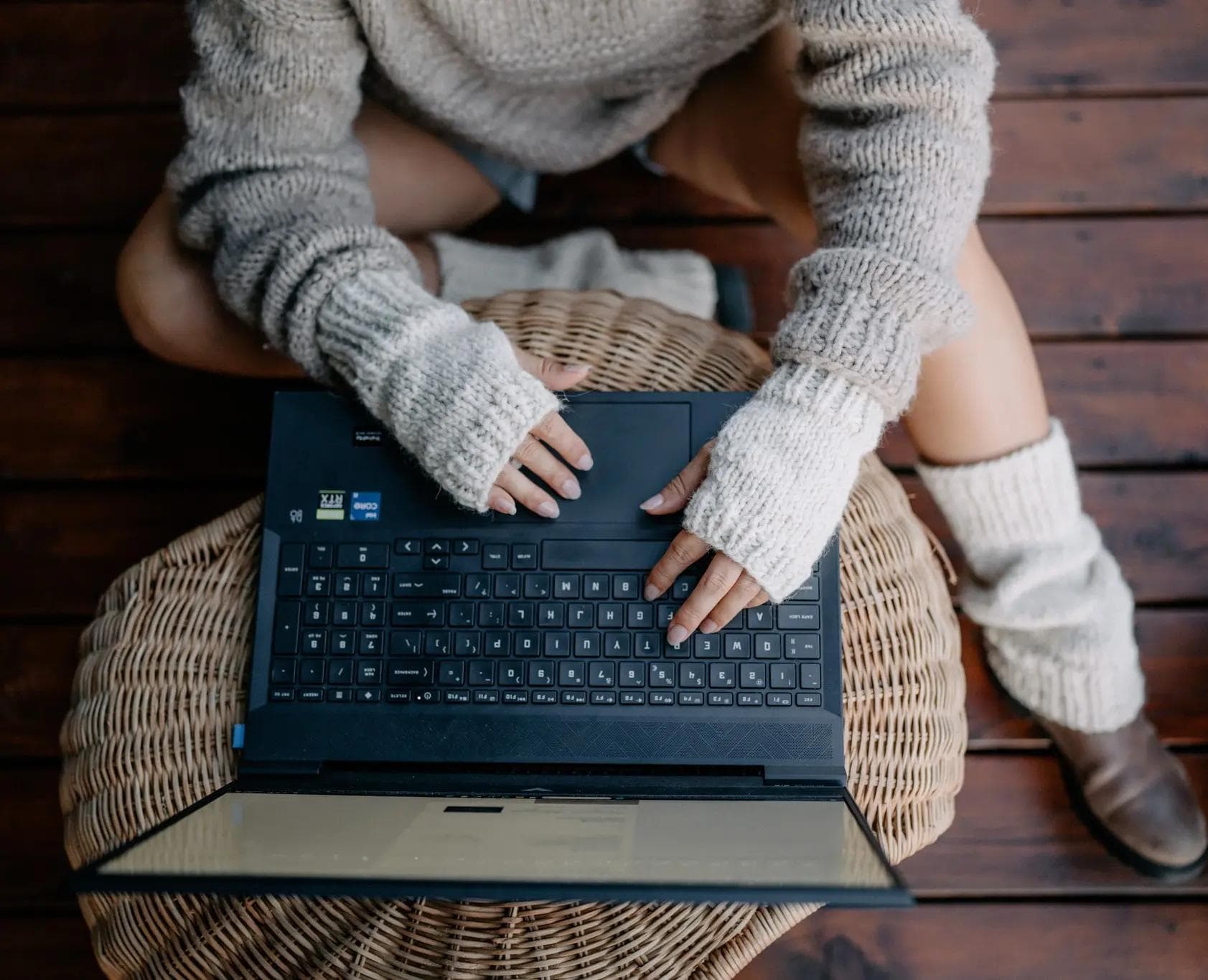 Zdjęcie przedstawia kobietę siedzącą na ciemnej, drewnianej podłodze. Ma na sobie gruby, beżowy sweter oraz ocieplacze na dłonie i nogi. Przed kobietą znajduje się otwarty laptop, który leży na wiklinowym pufie, a jej ręce spoczywają na klawiaturze. Zdjęcie jest zrobione z góry.