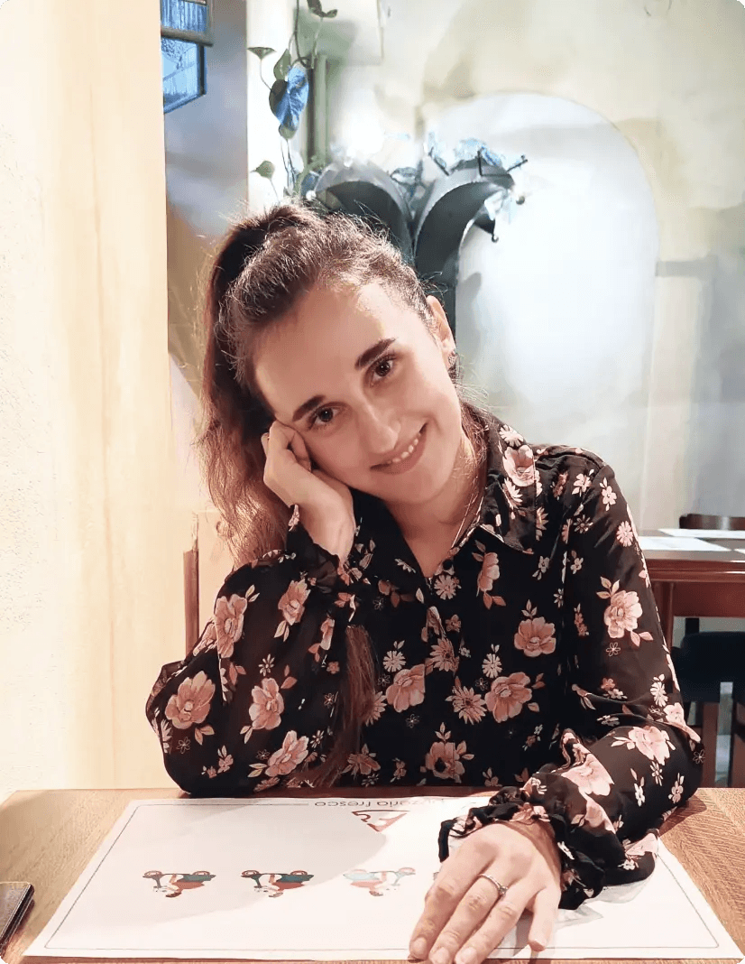 Na zdjęciu widzimy uśmiechniętą Magdalenę, która siedzi przy stole w restauracji lub kawiarni. Magda opiera głowę na dłoni, co nadaje zdjęciu spokojny i przyjazny charakter. Ma na sobie czarną bluzkę z różowym kwiatowym wzorem, a jej włosy związane są w kucyk. Na stole, przed nią znajduje się podkładka. W tle można dostrzec jasne ściany, roślinność i inne stoły z krzesłami.