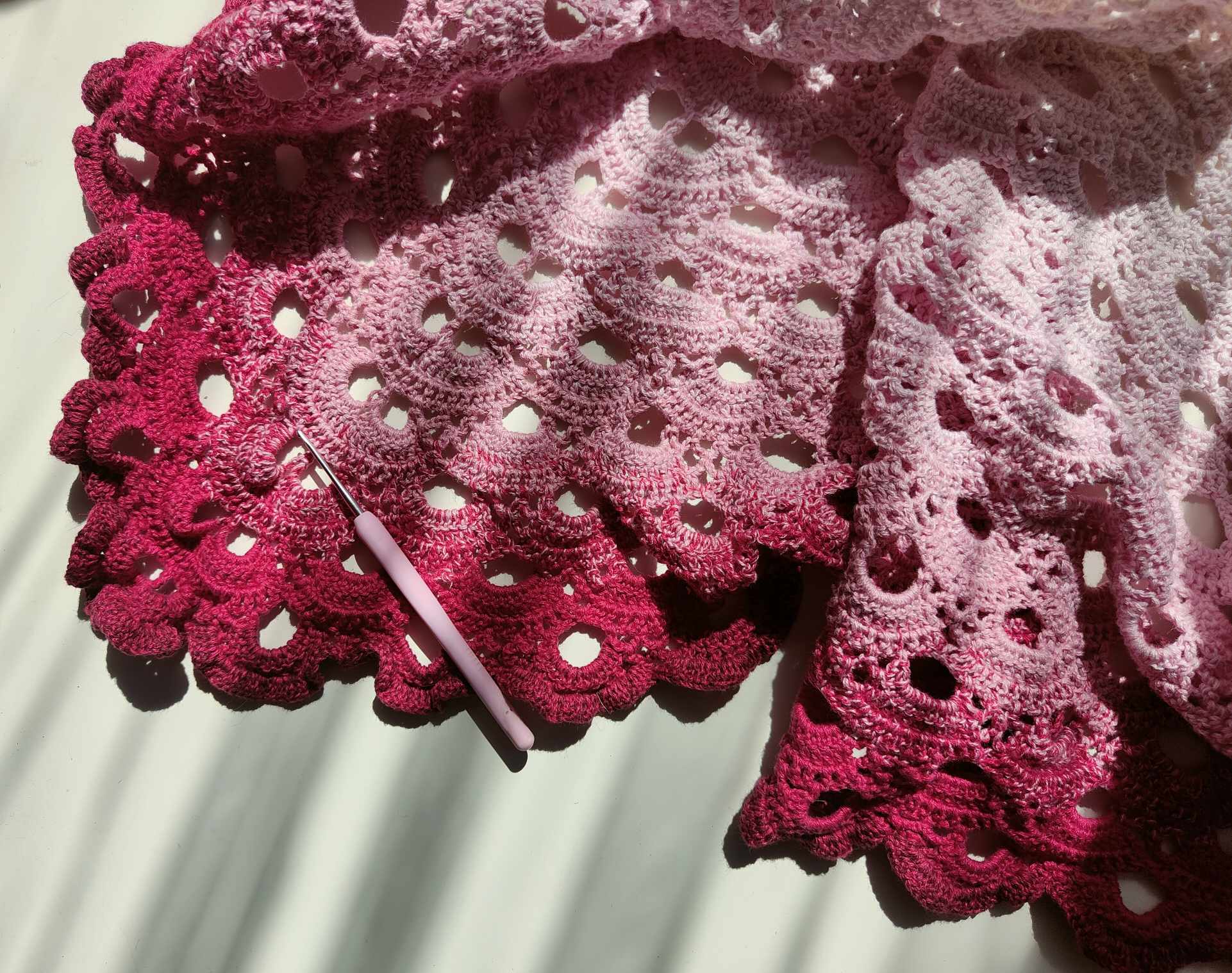 Obraz przedstawia projekt szydełkowy ze wzorem przechodzącym od ciemnoróżowego do jasnoróżowego. Szydełkowa praca ma falistą krawędź i ażurowy design z okrągłymi otworami. Na szydełkowej robótce znajduje się szydełko z różową rączką.