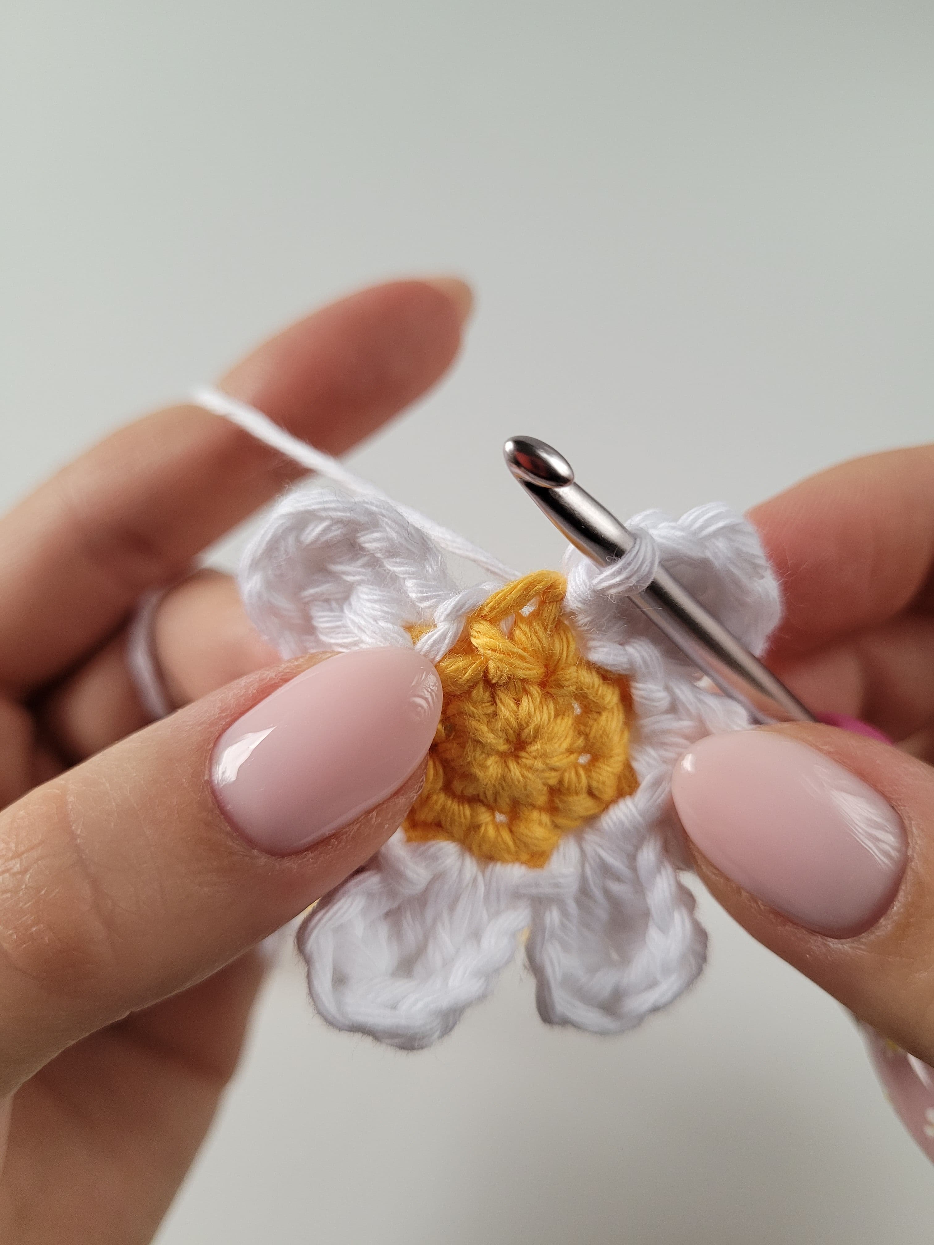 Obraz przedstawia zbliżenie na kobiece dłonie, które szydełkują mały kwiat. Kwiat ma żółty środek i białe płatki. Kobieta trzyma szydełko oraz włóczkę, pracując nad kwiatem. Jej paznokcie są pomalowane na jasnoróżowy kolor.