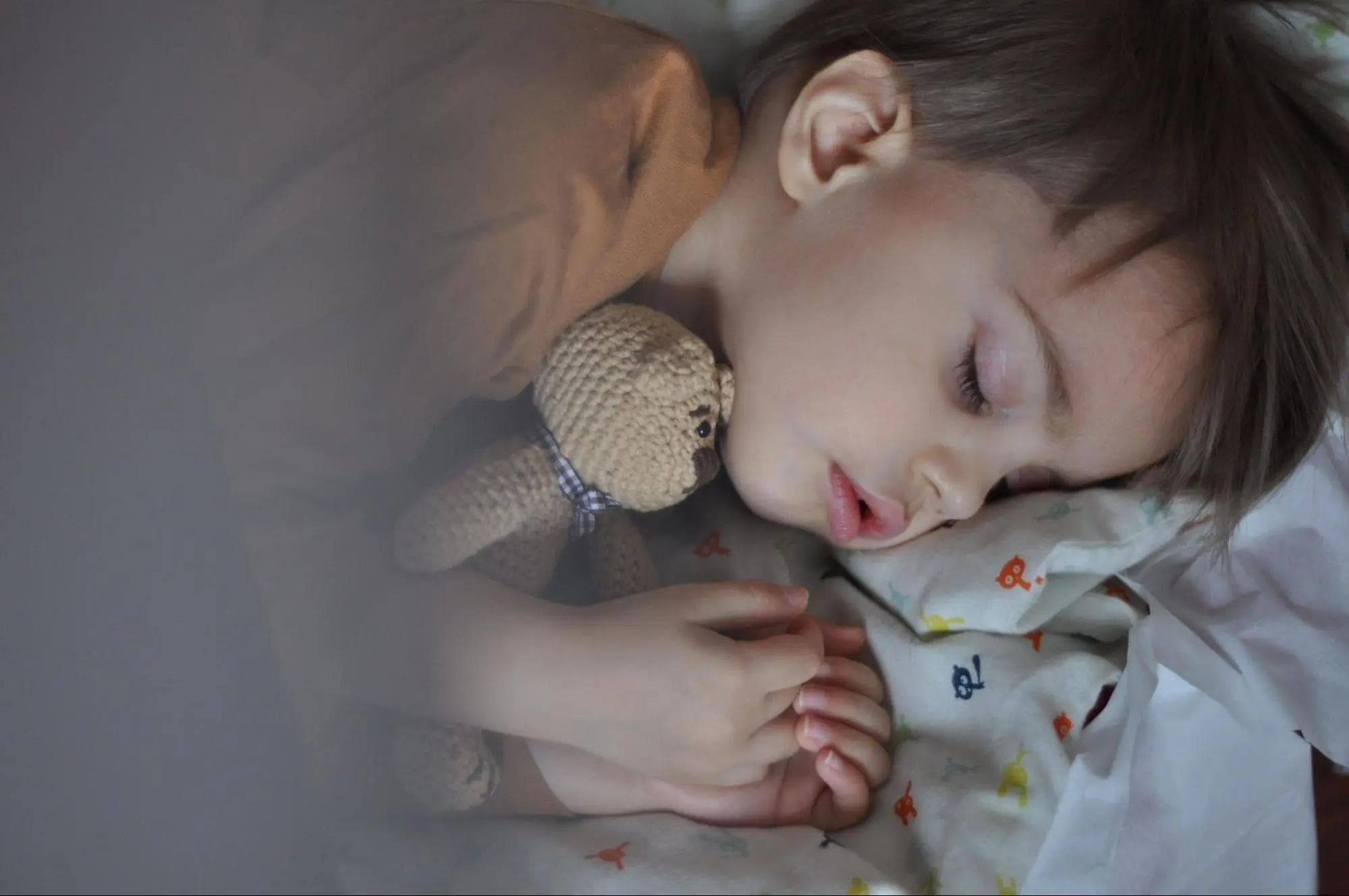 Na zdjęciu przedstawione śpiące dziecko, które przytula misia zrobionego na szydełku. Dziecko ma ubraną brązową koszulkę.