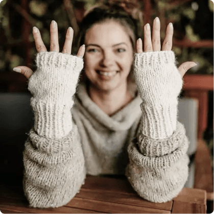 Na zdjęciu uśmiechnięta kobieta, która ma zamazaną twarz. Kobieta ma na sobie jasny, dziergany sweter i pasujące do niego rękawiczki bez palców. Osoba trzyma obie ręce w górze, prezentując rękawiczki. Tło jest rozmyte, a w tle widać trochę zieleni