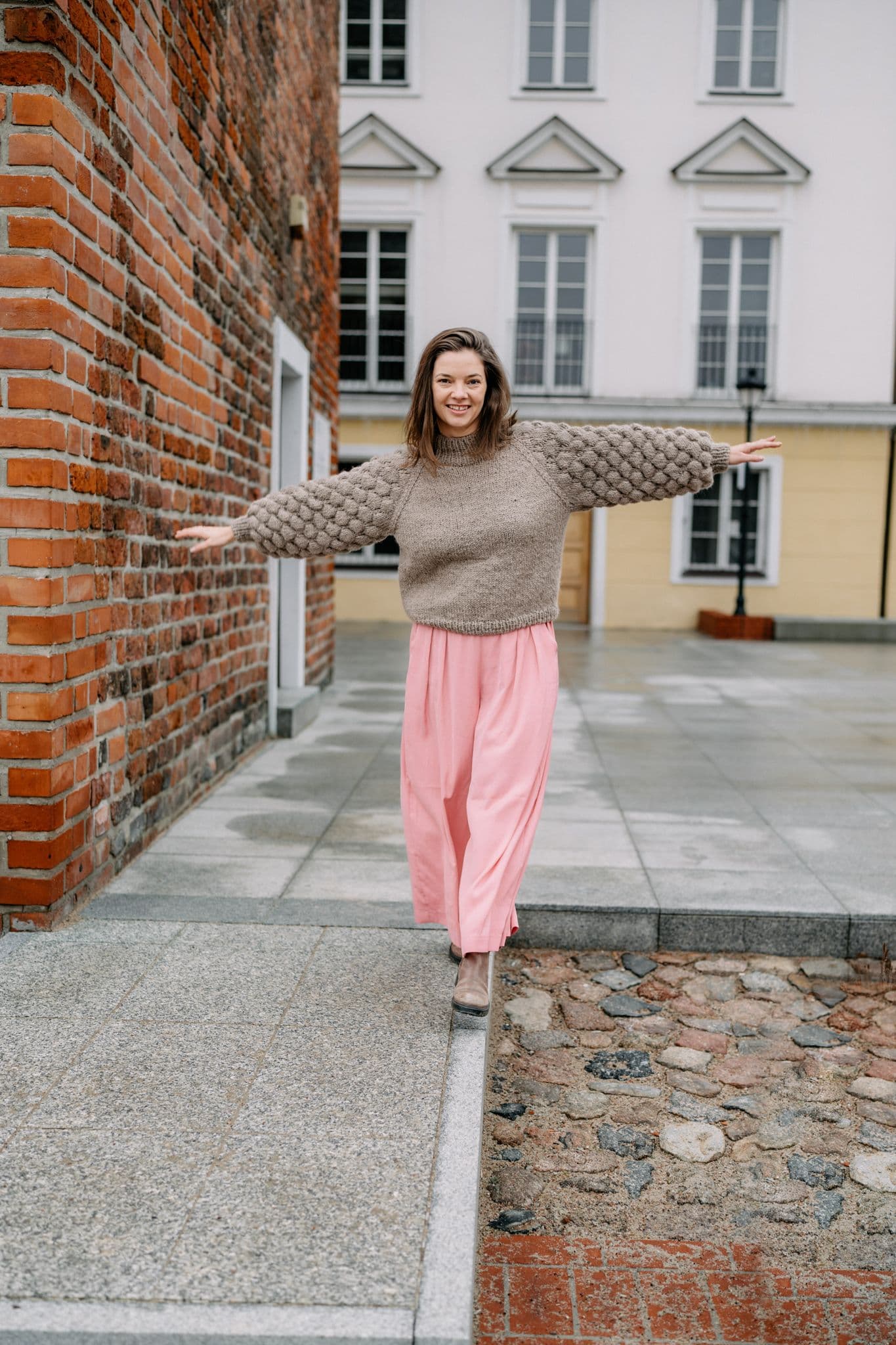 Zdjęcie przedstawia kobietę stojącą na krawędzi chodnika z rozłożonymi rękami. Kobieta ma na sobie brązowy sweter ze wzorem na rękawach i różowe spodnie. w tle znajduje się ceglana ściana po lewej stronie i budynek z białymi ścianami oraz wieloma oknami po prawej.