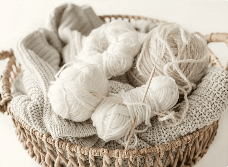 Na zdjęciu znajduje się kosz wypełniony włóczką i dzianiną. W koszu można zauważyć kilka kłębków włóczki w odcieniach bieli i beżu, a także druty do robienia na drutach. Włóczka jest ułożona na dzianinowych swetrach lub szalikach w podobnych kolorach.